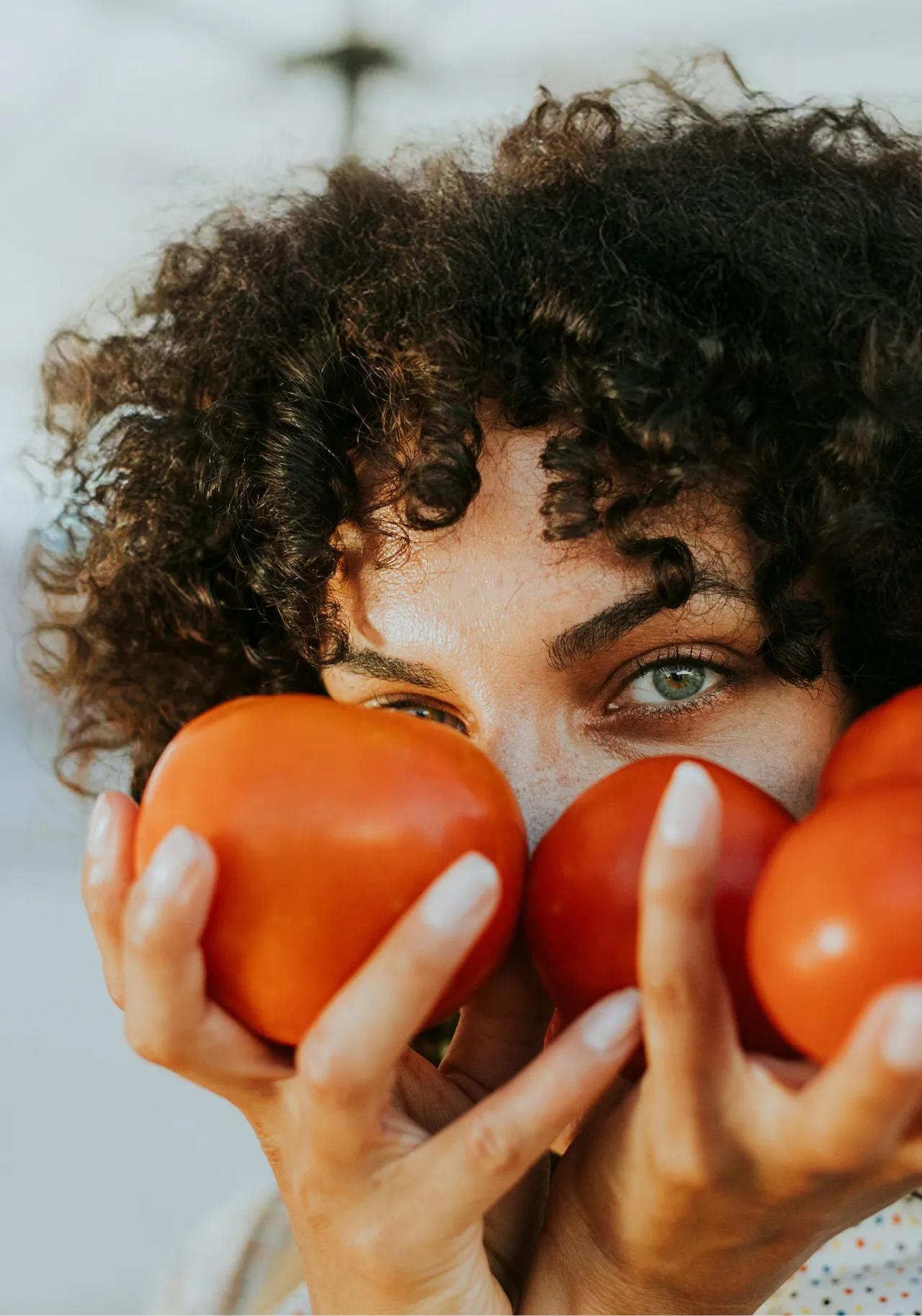 Frau mit lockigem Haar und Strohhut hält frische rote Tomaten vor ihr Gesicht. Symbol für gesunde Ernährung, Frische und nachhaltigen Lebensstil mit regionalem Gemüse und natürlichem Genuss.